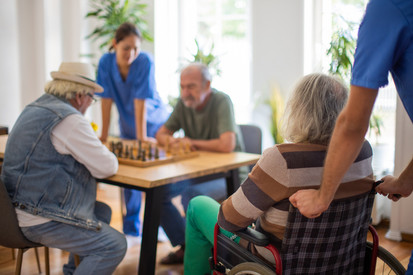 Pflegerin an einem Tisch, an dem zwei Männer Schach spielen, eine Frau wird mit dem Rollstuhl herangefahren