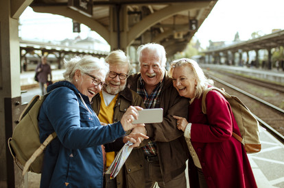 Vier Seniorinnen und Senioren stehen an einem Bahn-Gleis und schauen lachend auf ein Smartphone.