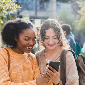 Zwei lächelnde Mädchen im Teenageralter lachen, während sie ein Video auf einem Smartphone ansehen. Sie stehen draußen auf dem Campus der Schule.