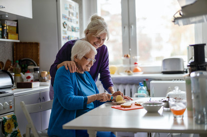 Eine ältere Frau sitzt an einem Küchentisch. Eine jüngere Frau steht hinter ihr und hält ihre Hand auf ihrer Schulter. 