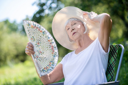 Eine alte Frau sitzt auf einem Gartenstuhl. Sie trägt einen großen Hut und hält einen Fächer.
