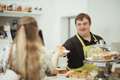 Ein Mann mit Down-Syndrom serviert einer Frau in einem Café einen Scone.