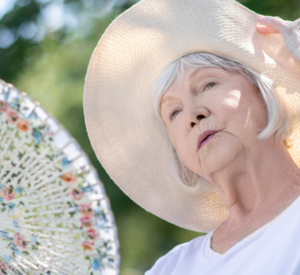 Eine alte Frau sitzt in einem Gartenstuhl im Schatten. Sie trägt einen großen Hut und hält einen Fächer.